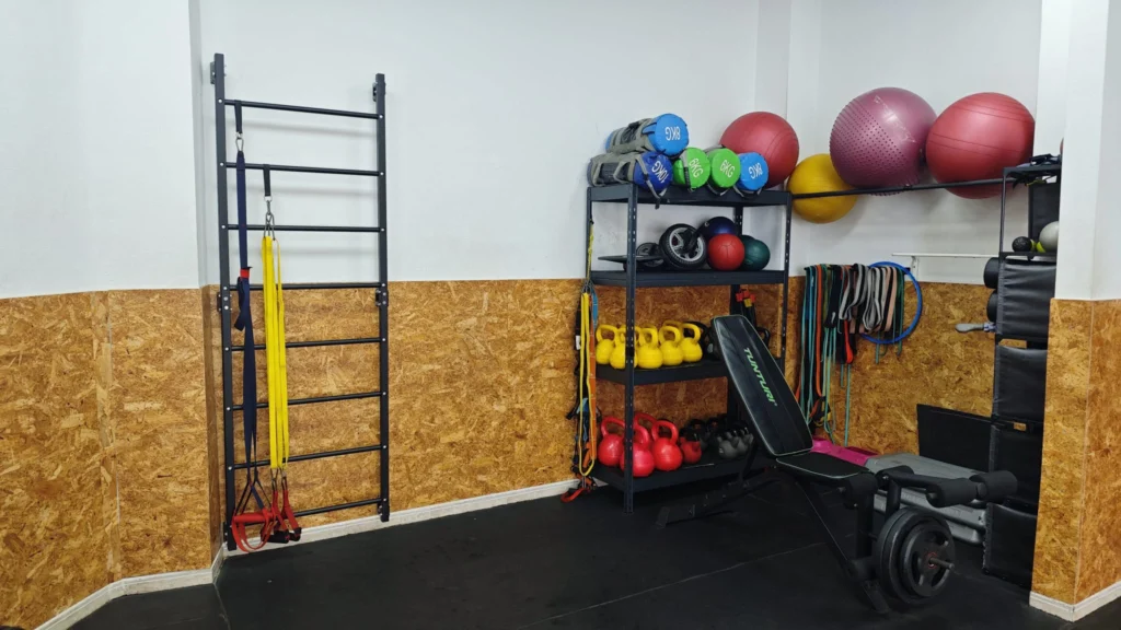Zona de entrenamiento funcional en un gimnasio, con espaldera de pared, cintas de suspensión, kettlebells, balones medicinales, bandas elásticas, fitballs y banco de pesas, organizada sobre estanterías metálicas y suelo de caucho negro.